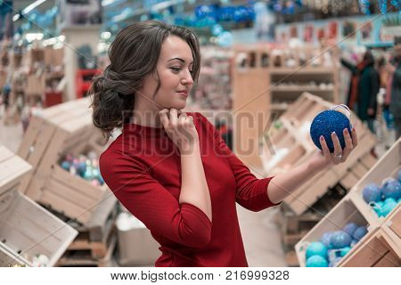 Cheerful happy customer girl choosing gifts for Christmas and New Year. She is taking Christmas tree decorations. difficult decision. Various choice