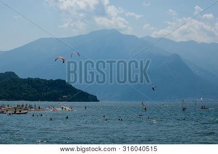 Lake Como, Italy - July 21, 2019: People Swimming In Mountain Lake. Kitesurfers And Windsurfers Trai