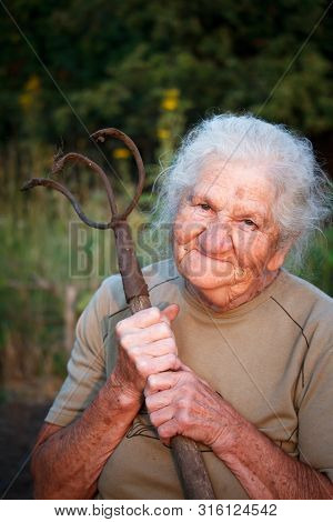 Close-up Portrait Of An Old Woman With Gray Hair Holding A Rusty Pitchfork Or Chopper In Her Hands,