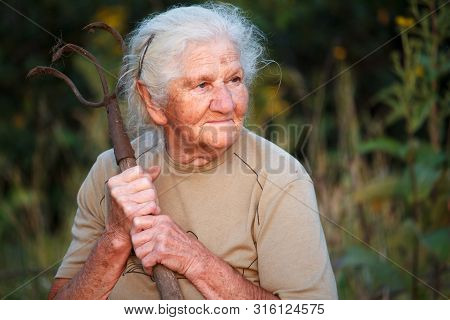 Close-up Portrait Of An Old Woman With Gray Hair Holding A Rusty Pitchfork Or Chopper In Her Hands,