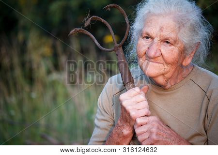 Close-up Portrait Of An Old Woman With Gray Hair Holding A Rusty Pitchfork Or Chopper In Her Hands,