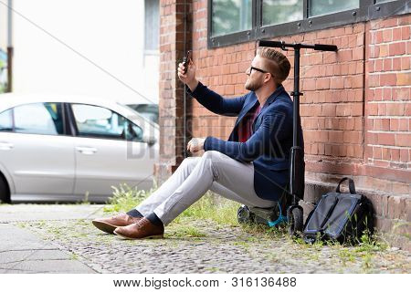Smiling Businessman Sitting On Electric Scooter Near Brick Wall Taking Selfie With Mobile Phone