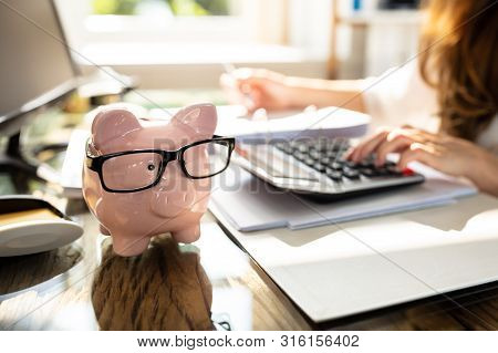 Businessperson Calculating Bill In Front Of Pink Piggy Bank Over Desk