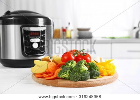 Wooden Board With Vegetables And Modern Multicooker On White Table In Kitchen. Space For Text