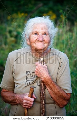 A Close-up Portrait Of An Old Woman With Gray Hair Smiling And Looking Up, Resting Her Chin On A Sti