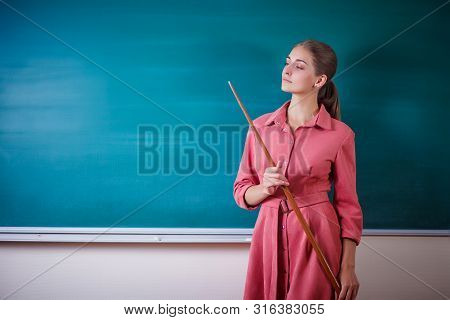 Young Woman Teacher Stands At A Blackboard With A Pointer. Template For The School Timetable And Oth