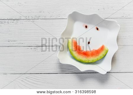 Cheerful Smiling Watermelon, A Face Made Of Slices And Bones On A White Plate. Wooden Background. Th