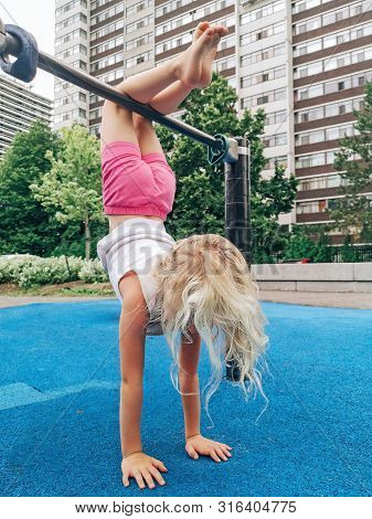 Cute Adorable Little Preschool Blonde Caucasian Girl Hanging Upside Down On Pullup Bar On Playground