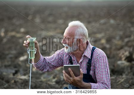 Senior Farmer With Tablet Squatting Beside Rain Gauge In Field