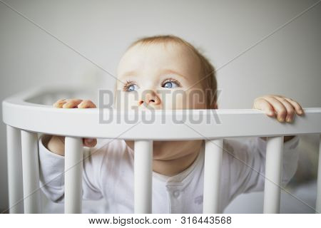 Adorable Baby Girl In Co-sleeper Crib Attached To Parents Bed. Little Kid Sitting In Cot