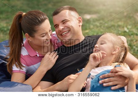 Young Happy Family Spending Free Time In Park On Picnic, Mother And Father Laugh From Doughter's Jok
