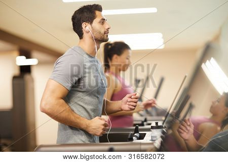 Side View Portrait Of Handsome Middle-eastern Man Running On Treadmill In Gym And Listening To Music