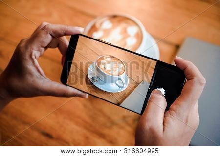 Hand Taking A Picture Of Hot Latte Art Coffee Cup With Art Foam On Wooden Table With Her Smart Phone