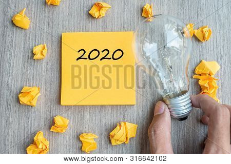 2020  Words On Yellow Note And Crumbled Paper With Businessman Holding Lightbulb On Wooden Table Bac