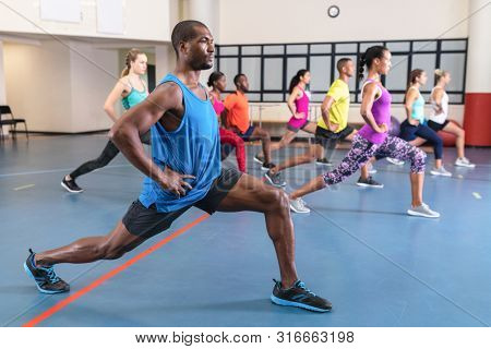Side view of African-american male trainer training people to perform yoga in fitness center. Bright modern gym with fit healthy people working out and training