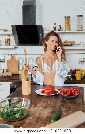 Smiling Woman In White Bra Talking Smartphone While Cooking In Kitchen