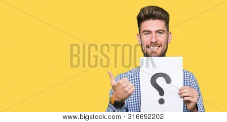 Young handsome man holding paper with question mark happy with big smile doing ok sign, thumb up with fingers, excellent sign