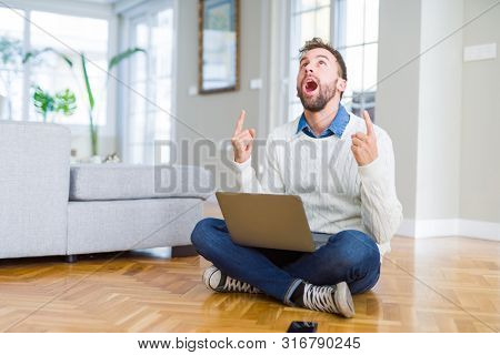 Handsome man wearing working using computer laptop amazed and surprised looking up and pointing with fingers and raised arms.