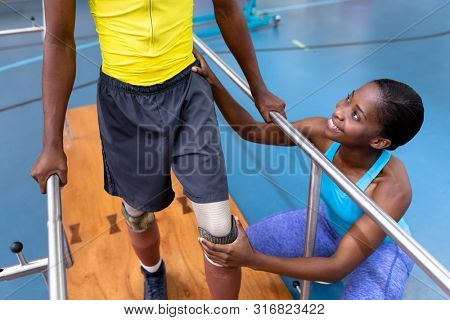 High angle view of African-american physiotherapist assisting disabled African-american man walk with parallel bars in sports center. Sports Rehab Centre with physiotherapists and patients working