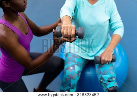 Close-up of African-american female trainer assisting disabled Caucasian senior woman to exercise with dumbbell in sports center. Sports Rehab Centre with physiotherapists and patients working