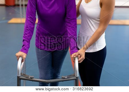 Mid section of female physiotherapist helping disabled senior woman walk with walker in sports center. Sports Rehab Centre with physiotherapists and patients working together towards healing