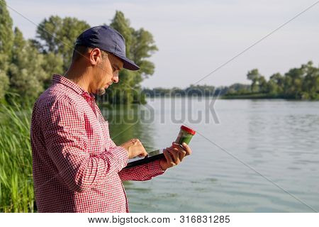 Side View Of Ecologist On River Bank Entering Data On Green Algae On Tablet