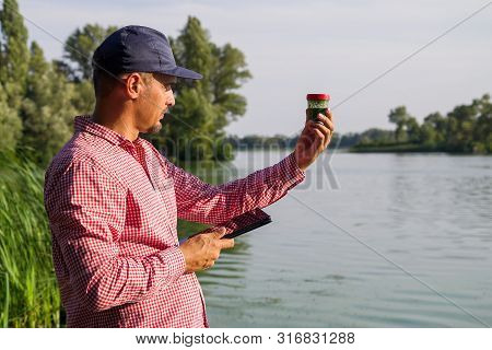 Ecologist On River Bank Examines Container With Green Algae And Holds Tablet