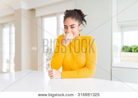 Beautiful african american woman with afro hair wearing a casual yellow sweater smelling something stinky and disgusting, intolerable smell, holding breath with fingers on nose. Bad smells concept.