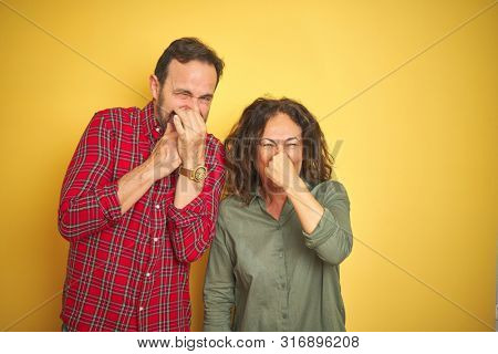 Beautiful middle age couple over isolated yellow background smelling something stinky and disgusting, intolerable smell, holding breath with fingers on nose. Bad smells concept.