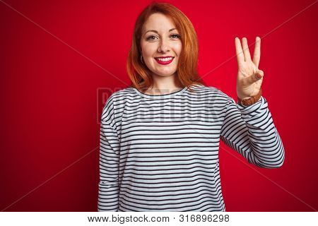 Young redhead woman wearing strapes navy shirt standing over red isolated background showing and pointing up with fingers number three while smiling confident and happy.