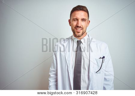 Young handsome doctor man wearing white profressional coat over isolated background winking looking at the camera with sexy expression, cheerful and happy face.