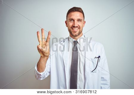 Young handsome doctor man wearing white profressional coat over isolated background showing and pointing up with fingers number three while smiling confident and happy.