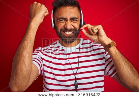 Young indian call center agent man using headset over isolated red background annoyed and frustrated shouting with anger, crazy and yelling with raised hand, anger concept