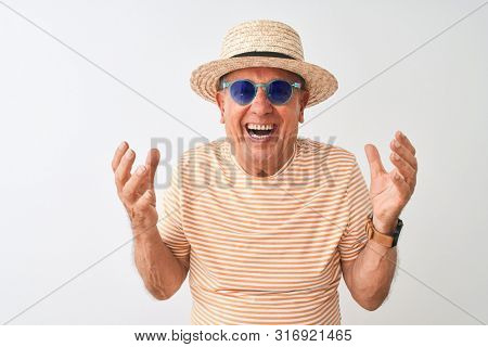 Senior man wearing striped t-shirt sunglasses and sumer hat over isolated white background very happy and excited, winner expression celebrating victory screaming with big smile and raised hands