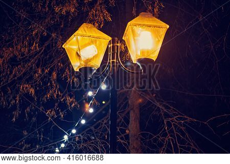 Two Old Street Light And Decorative Outdoor String Lights Hanging On Tree At Night Time. Yellow Lant