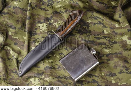 A Knife With A Wooden Handle In A Leather Sheath And A Metal Flask On Camouflage Cloth. A Large Knif
