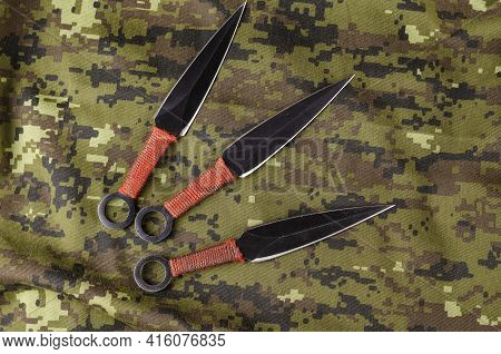 Black Throwing Knives On Camouflage Cloth. Three Special Knives With Red Handles Crossed. Military T