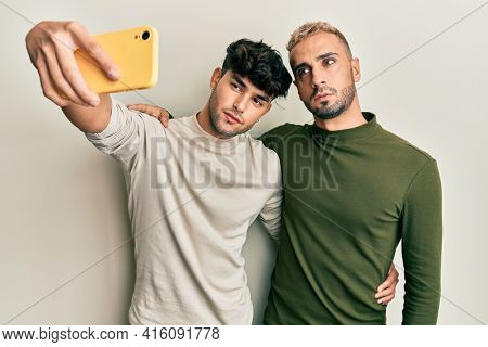 Homosexual gay couple standing together taking a selfie photo with smartphone thinking attitude and sober expression looking self confident 