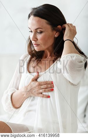 Middle aged woman sitting on a white bed in a bedroom at home touching chest with her hands while having a heart pain and feeling unwell. Suffering from stress, hypertension or cardiac arrhythmia.
