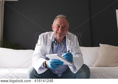 Smiling caucasian senior male doctor making notes during consultation video call. telemedicine online during quarantine lockdown.