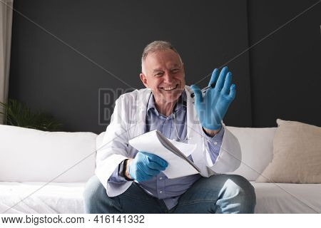 Smiling caucasian senior male doctor holding notebook waving during consultation video call. telemedicine online during quarantine lockdown.