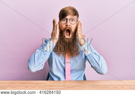Young irish redhead man wearing business shirt and tie sitting on the table afraid and shocked, surprise and amazed expression with hands on face 
