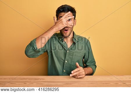 Young handsome man wearing casual clothes sitting on the table peeking in shock covering face and eyes with hand, looking through fingers afraid 