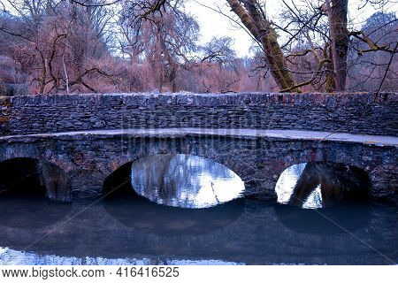 Old Stone Footbridge Over The River Bybrook With Reflection Of The Woodland With Romantic Colours In