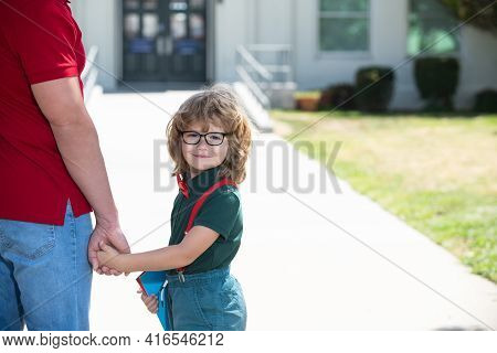 Father And Son Walking Trough School Park. Portrait Of Happy Nerd Pupil Holding Teachers Hand