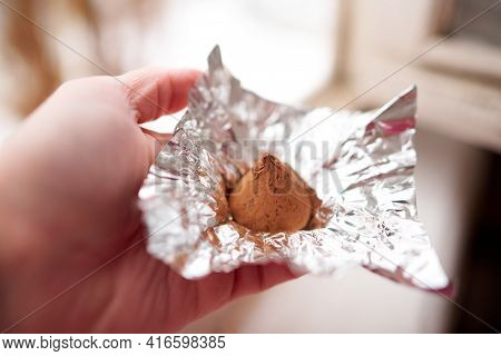 A Female Hand Holds A Chocolate Truffle Candy Wrapped In A Foil Wrapper