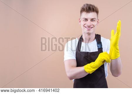 Young Smiling Man Wearing Cleaner Apron And Rubber Yellow Gloves Isolated On Beige Background. Happy