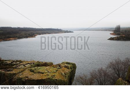 At The Bend Of The River There Is A Large Rock, Trees In The Distance