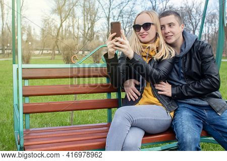 Husband And Wife Take A Selfie While Sitting On A Swing In The Park. Happy Couple Taking Pictures Of