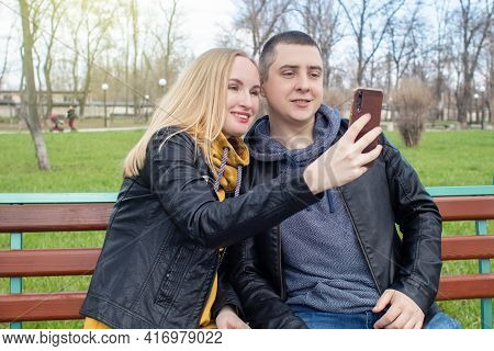 Husband And Wife Take A Selfie While Sitting On A Swing In The Park. Happy Couple Taking Pictures Of